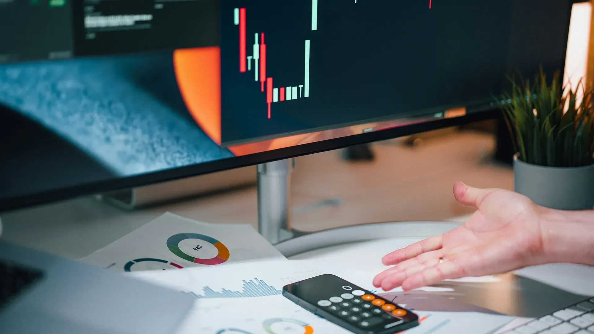 A hand reaching for a calculator beside printed financial charts, with trading charts visible on a monitor behind