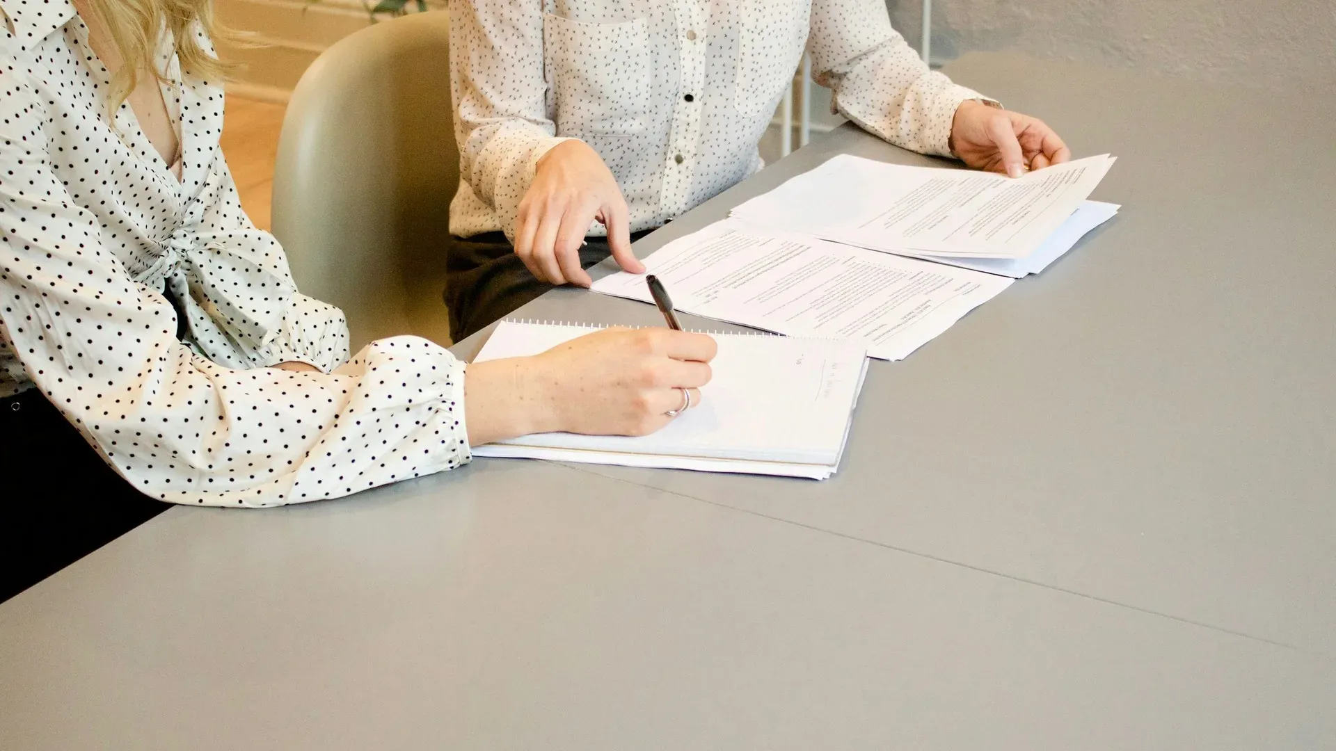 Two people at an office table reviewing printed documents, one writing in a notebook, faces out of frame
