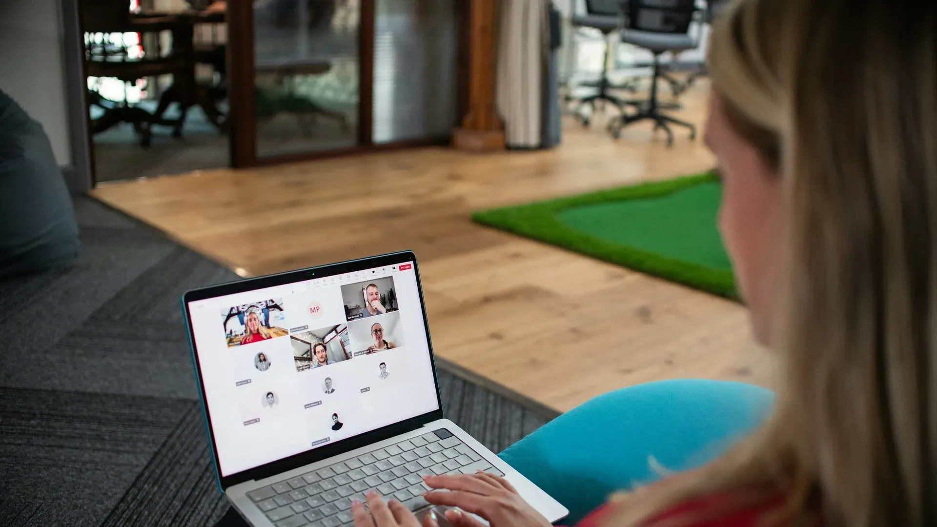 Over-the-shoulder view of a woman on a laptop video call showing a grid of multiple participant tiles, in an open-plan office