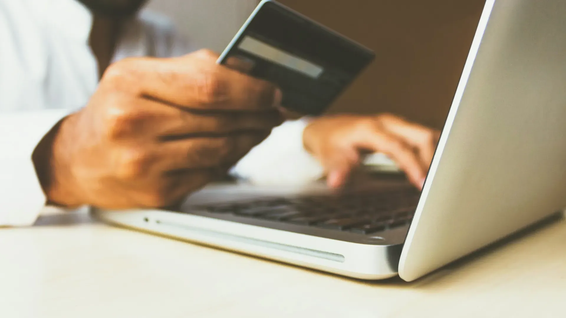 A hand holding a bank card above a laptop keyboard, with the screen out of focus behind it