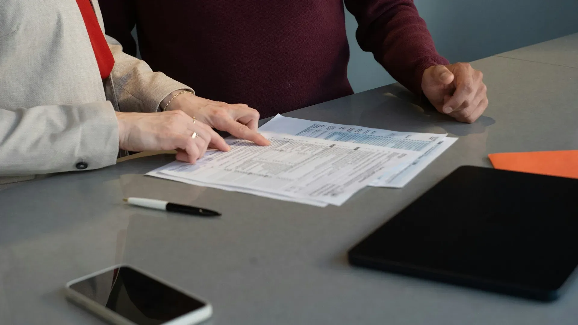 Two people at an office desk reviewing a printed form together, one pointing at a line item, with a phone and tablet on the table