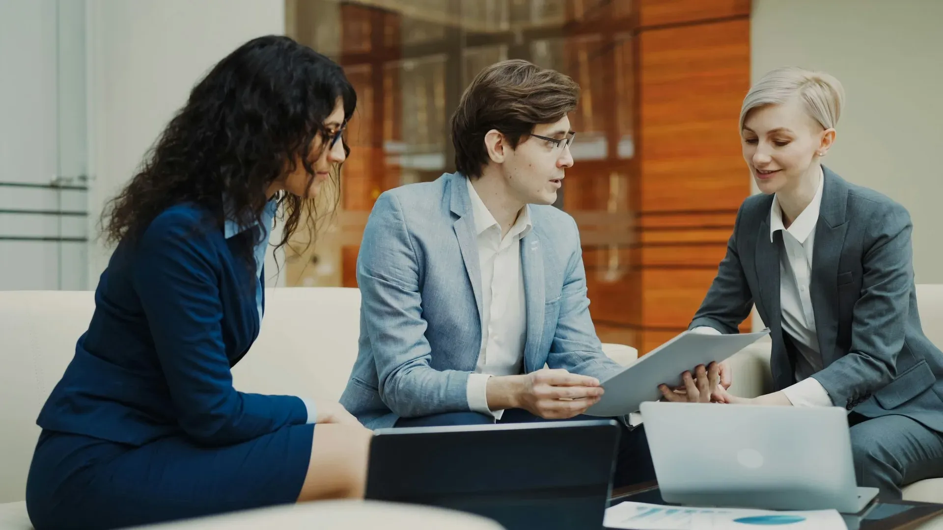 Three business professionals in an office lounge discussing a printed document together, with two laptops open on the table in front of them