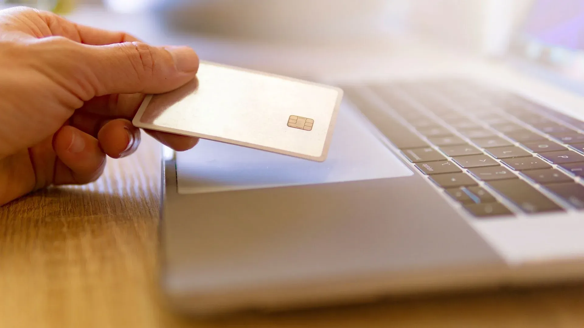A hand holding a blank bank card with a visible chip beside an open laptop keyboard on a wooden desk