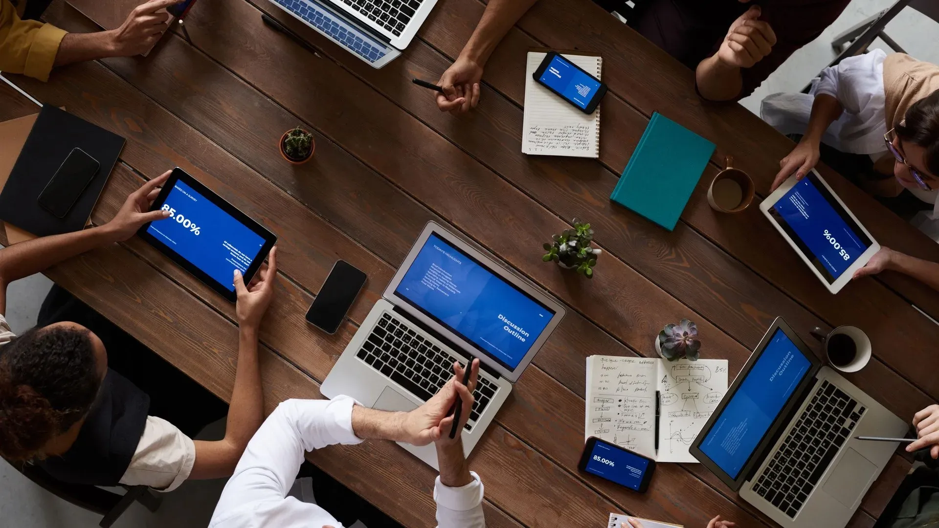 Overhead view of a team meeting around a wooden table with laptops and tablets showing blue data dashboard screens, notebooks and coffee cups
