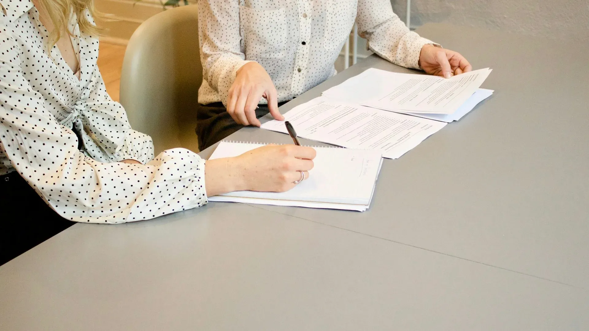 Two people at an office desk reviewing paper documents, one signing a notebook page with a pen while the other holds a printed document