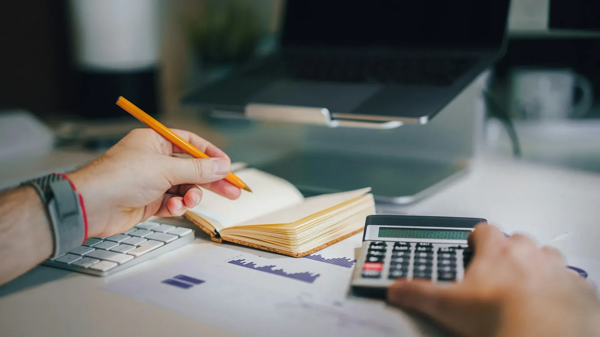 Close-up of hands holding a pencil over an open notebook next to a calculator and a printed bar chart, with a laptop in the background