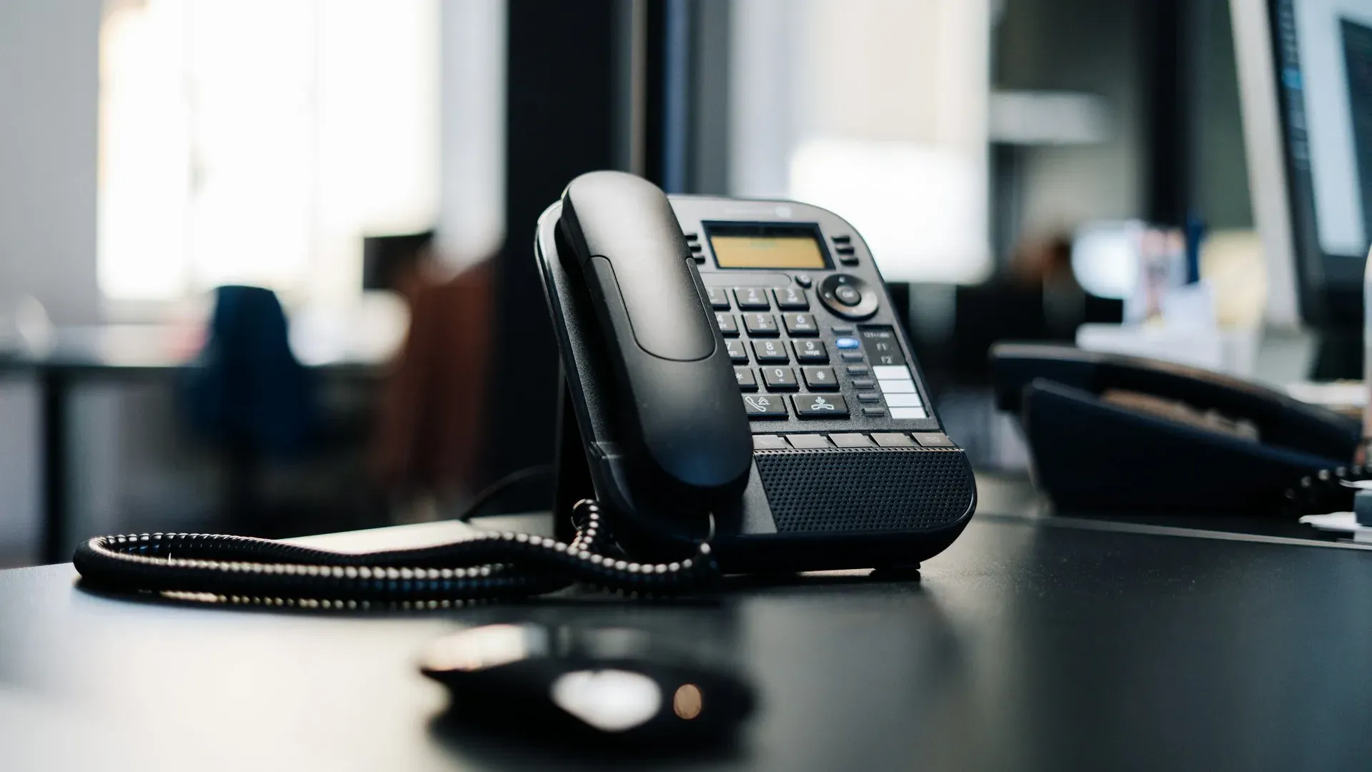 A black office desk phone with a coiled cord, blurred office workspace in the background