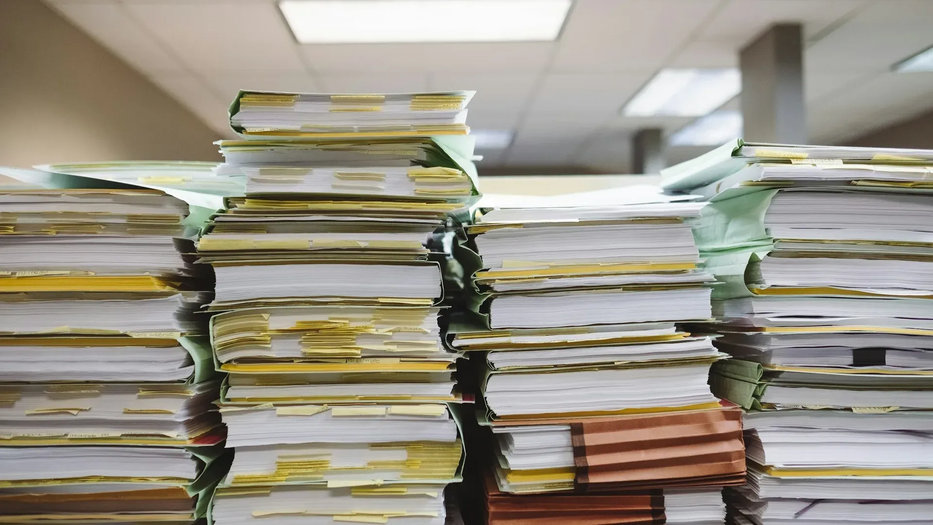 Tall stacks of paper files and folders on an office desk under fluorescent lighting