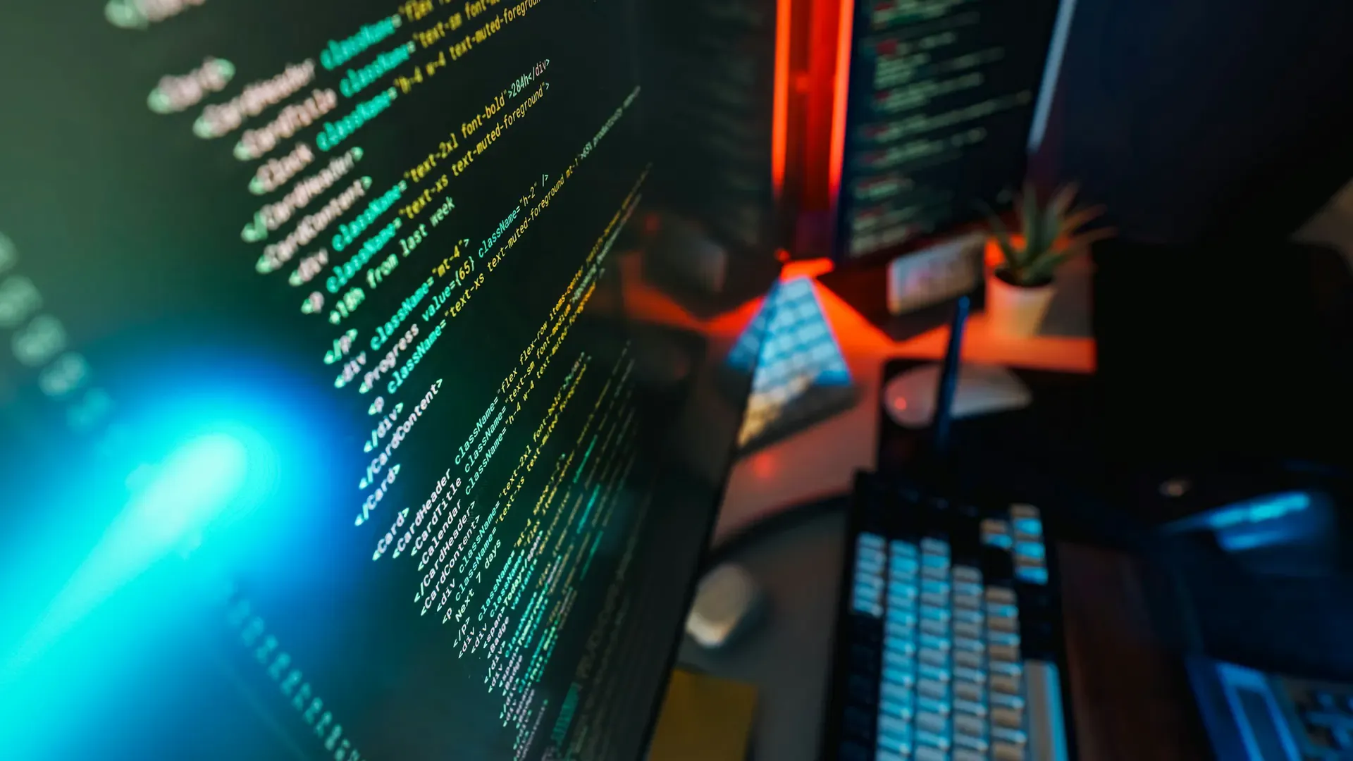 A computer monitor in a dark room showing lines of colourful code, with a lit keyboard and desk setup beside it