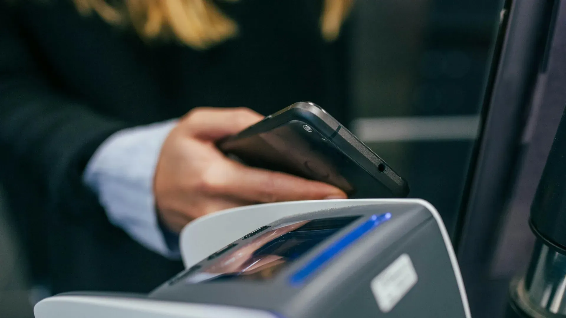 Extreme close-up of a hand tapping a smartphone against a payment terminal with a blue accent light, business suit sleeve visible