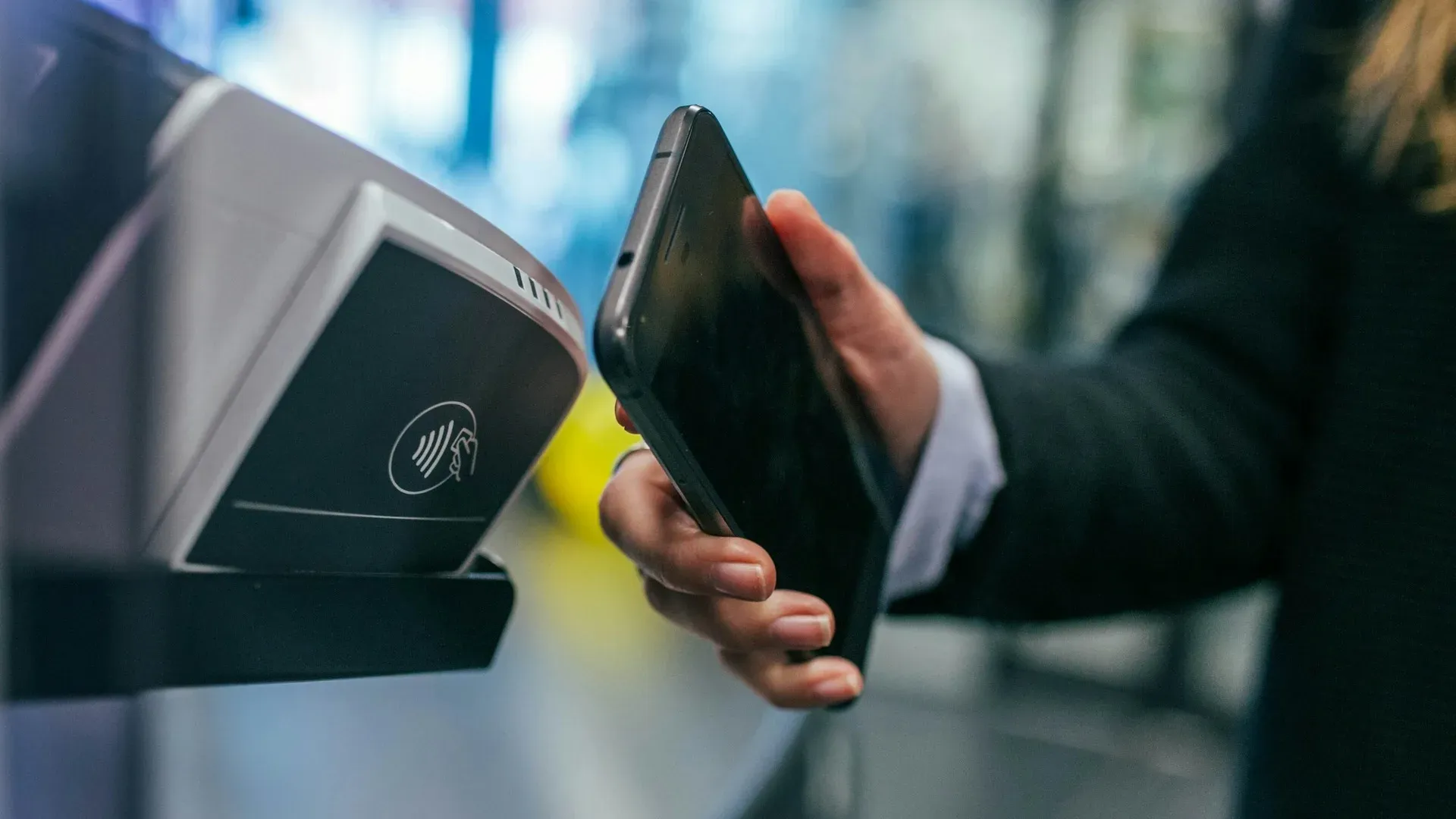 A businessperson's hand holding a smartphone up to tap on a contactless payment terminal, with a blurred retail interior in the background