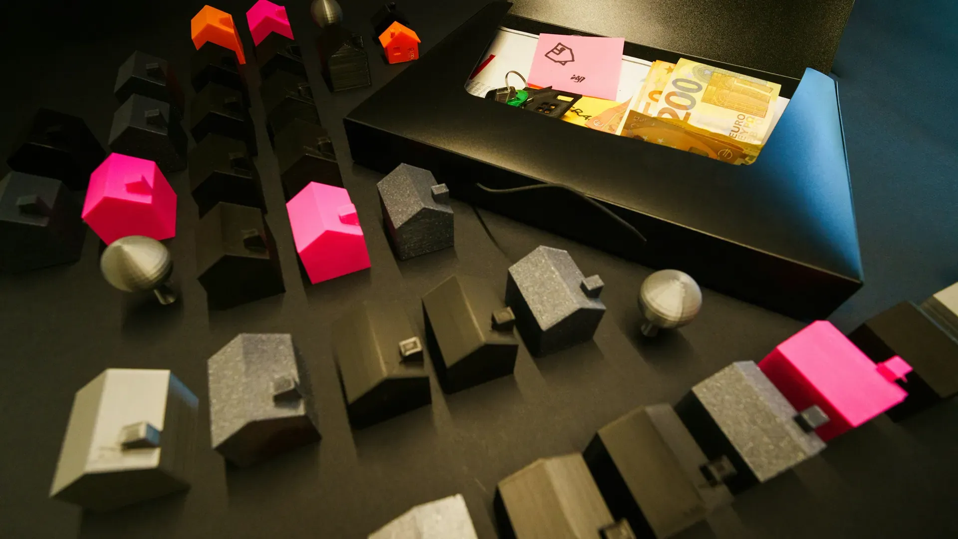 Small model houses arranged on a dark surface beside an open box holding banknotes, keys and a note
