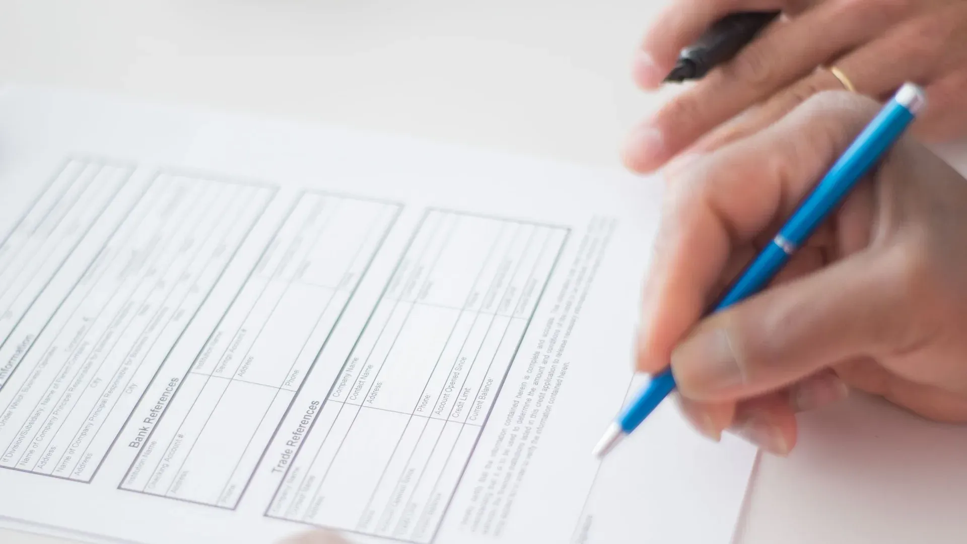 Two people's hands over a bank references and trade references form, one hand holding a blue pen ready to sign