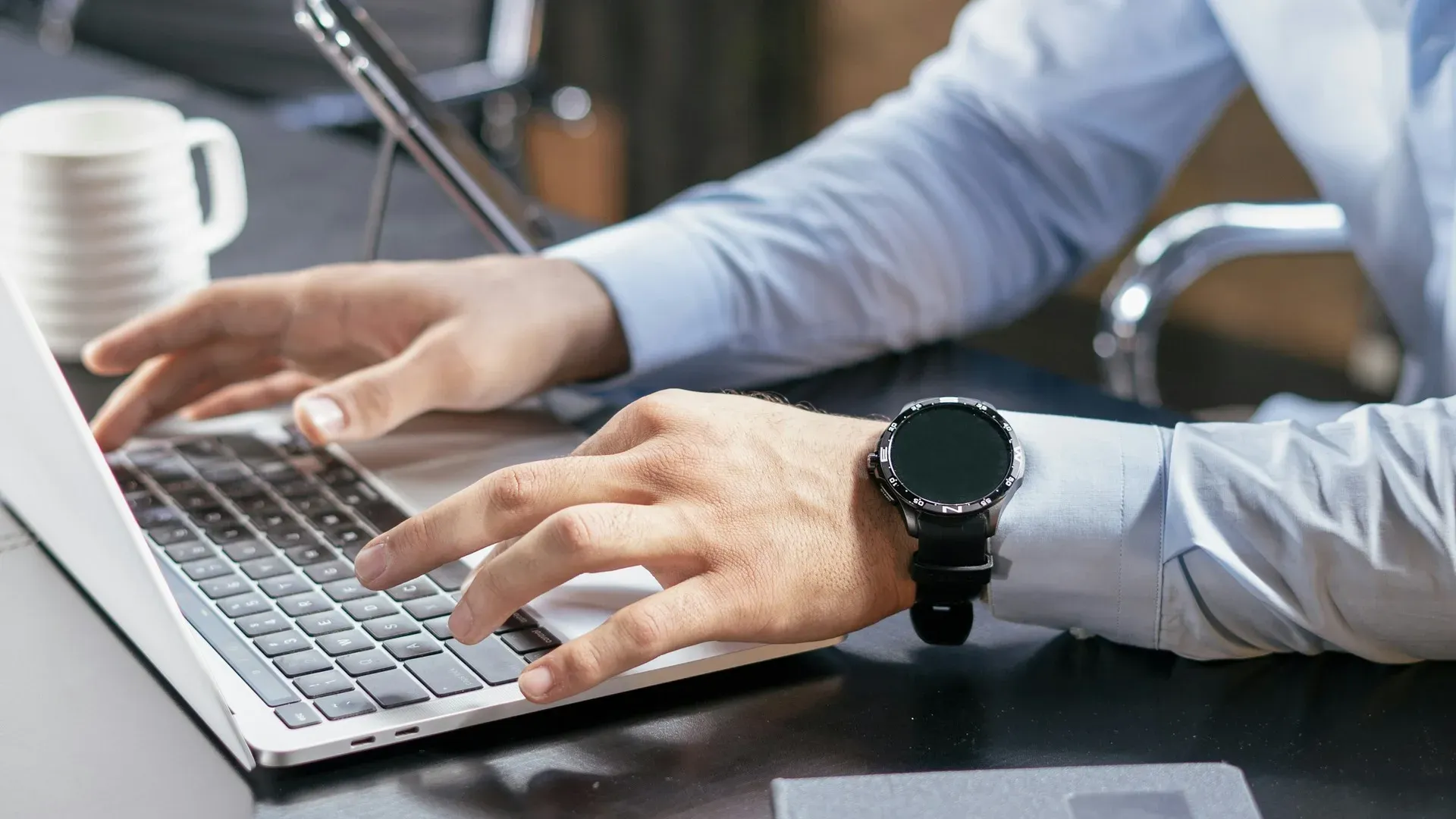 Close-up of hands typing on a laptop keyboard at a desk, with a coffee mug and smartwatch visible